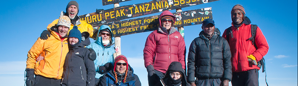 A group of people taking a picture of a sign at the top of Mount Kilimanjaro